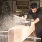 Un falegname usa una piallatrice per levigare un grosso tronco di legno, creando una nuvola di segatura. Il piallare significa rendere liscio e uniforme.