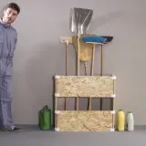 Un uomo in tuta grigia accanto a un porta attrezzi da giardino fai da te, con scope, palette e annaffiatoio.