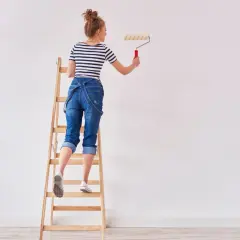 Donna su scala per pitturare casa fai da te, con rullo in mano.