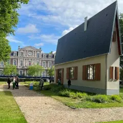 La casa più piccola del mondo, un'abitazione compatta con tetto spiovente, si erge in un parco con un edificio storico sullo sfondo.