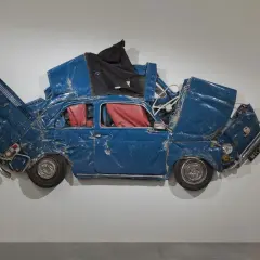 Opera di Ron Arad: una Fiat 500 blu, schiacciata e deformata, con interni rossi, appesa a un muro.