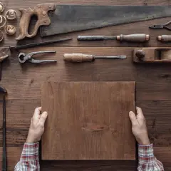 Mani che tengono un pezzo di legno, circondate da attrezzi da falegname, mostrando come si lavora il legno.
