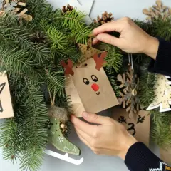 Mani che decorano un calendario dell'avvento con sacchetti natalizi fai da te, pigne e decorazioni in legno.