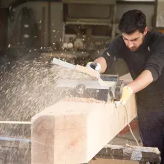 Un falegname usa una piallatrice per levigare un grosso tronco di legno, creando una nuvola di segatura. Il piallare significa rendere liscio e uniforme.
