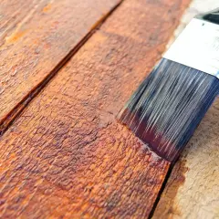Pennello stende impregnante su assi di legno grezzo, creando una finitura protettiva e colorata.