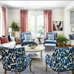 Salotto in stile grandmillennial con divano bianco, poltrone blu e sedie con fantasia floreale blu e bianca. Tende rosse e specchio dorato.