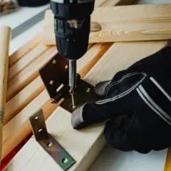 Mano guantata usa un avvitatore per unire due pezzi di legno con una staffa angolare e viti, mostrando come unire due pezzi di legno con viti.
