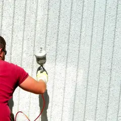 Uomo con maglietta rossa effettua verniciatura a spruzzo fai da te su una parete esterna bianca e rigata.