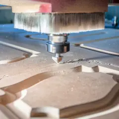 La fresa CNC sta lavorando il legno, creando un intricato motivo decorativo. La polvere di legno vola mentre la macchina esegue il suo lavoro di precisione.