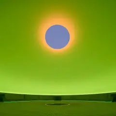 Interno di un'installazione di James Turrell: un cerchio blu su sfondo verde brillante, con un alone arancione. La sala circolare ha sedute e un cerchio luminoso al centro.