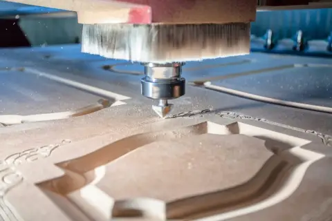 La fresa CNC sta lavorando il legno, creando un intricato motivo decorativo. La polvere di legno vola mentre la macchina esegue il suo lavoro di precisione.