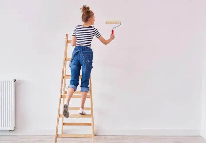 Donna su scala per pitturare casa fai da te, con rullo in mano.