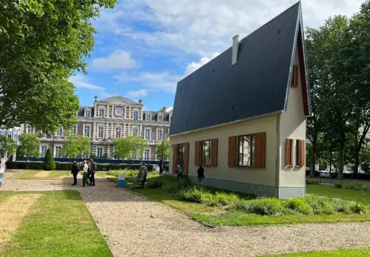 La casa più piccola del mondo, un'abitazione compatta con tetto spiovente, si erge in un parco con un edificio storico sullo sfondo.