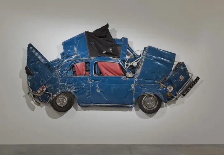 Opera di Ron Arad: una Fiat 500 blu, schiacciata e deformata, con interni rossi, appesa a un muro.
