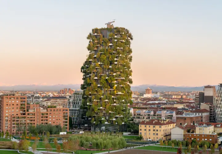 Il Bosco Verticale di Milano, un grattacielo ricoperto di alberi e piante, si staglia contro un cielo al tramonto, circondato da altri edifici della città.
