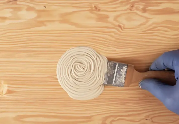 Mano con guanto blu applica stucco con pennello su legno. Guida pratica su come stuccare il legno per una finitura perfetta.