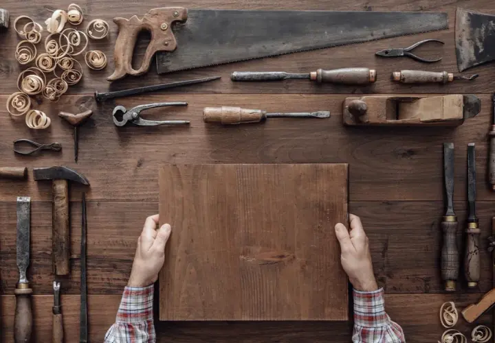Mani che tengono un pezzo di legno, circondate da attrezzi da falegname, mostrando come si lavora il legno.