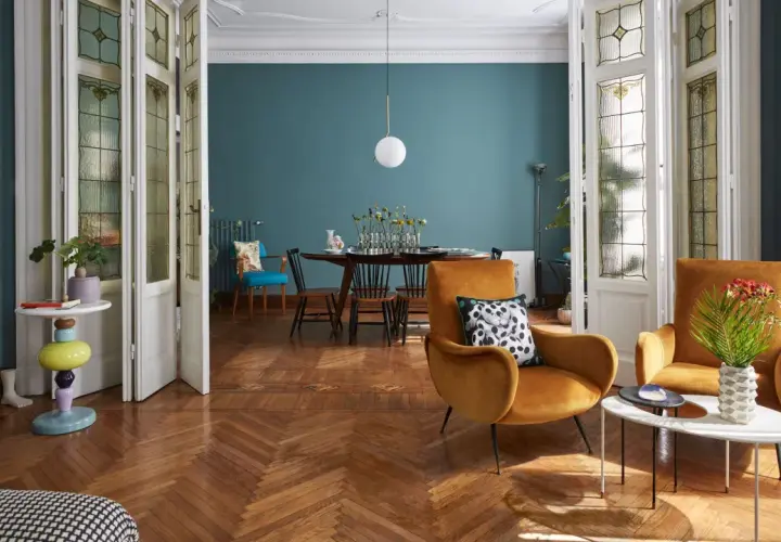 Soggiorno con poltrone ocra, tavolino moderno e un tavolo da pranzo in stile direttorio sullo sfondo. Porte a vetri decorati.
