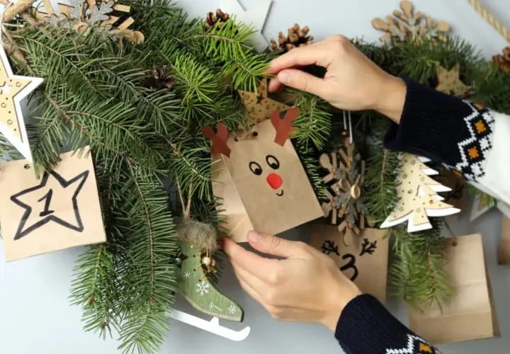 Mani che decorano un calendario dell'avvento con sacchetti natalizi fai da te, pigne e decorazioni in legno.