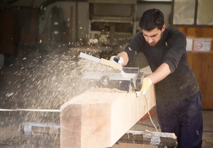 Un falegname usa una piallatrice per levigare un grosso tronco di legno, creando una nuvola di segatura. Il piallare significa rendere liscio e uniforme.