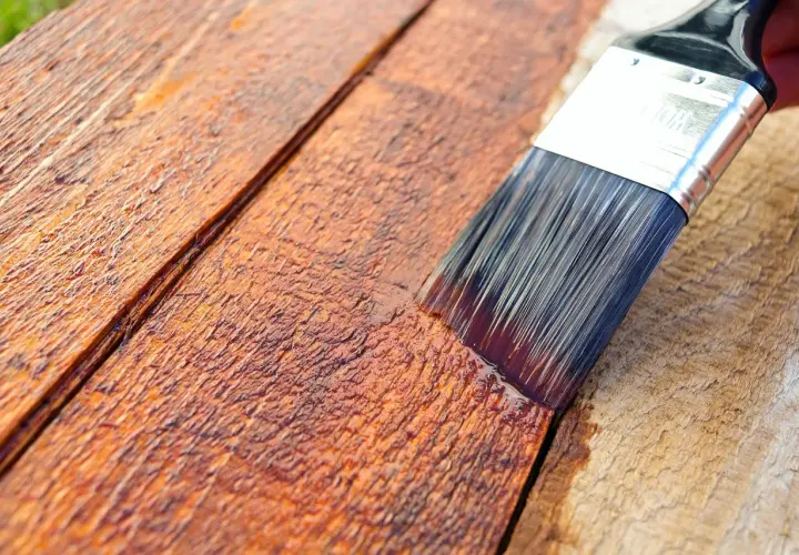 Pennello stende impregnante su assi di legno grezzo, creando una finitura protettiva e colorata.
