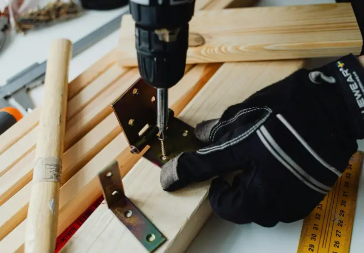 Mano guantata usa un avvitatore per unire due pezzi di legno con una staffa angolare e viti, mostrando come unire due pezzi di legno con viti.