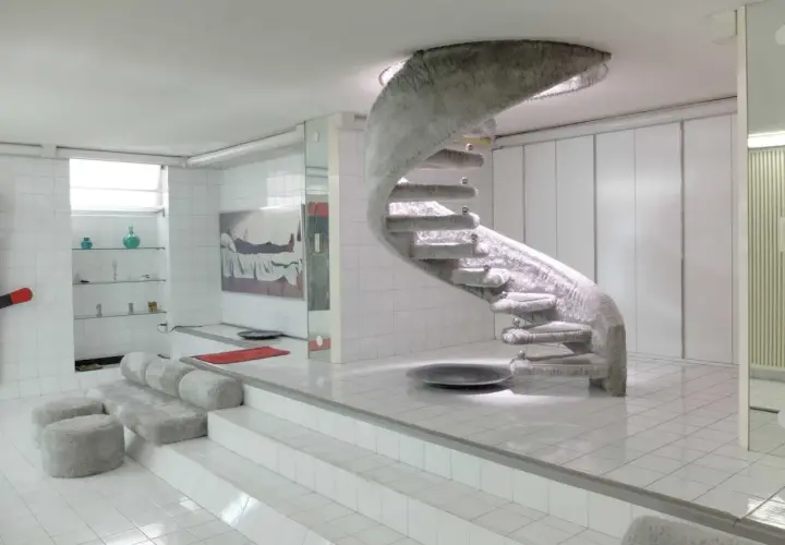 Interni futuristici con scala a chiocciola rivestita in pelliccia, un omaggio a Nanda Vigo. Divani e pouf morbidi completano l'ambiente.
