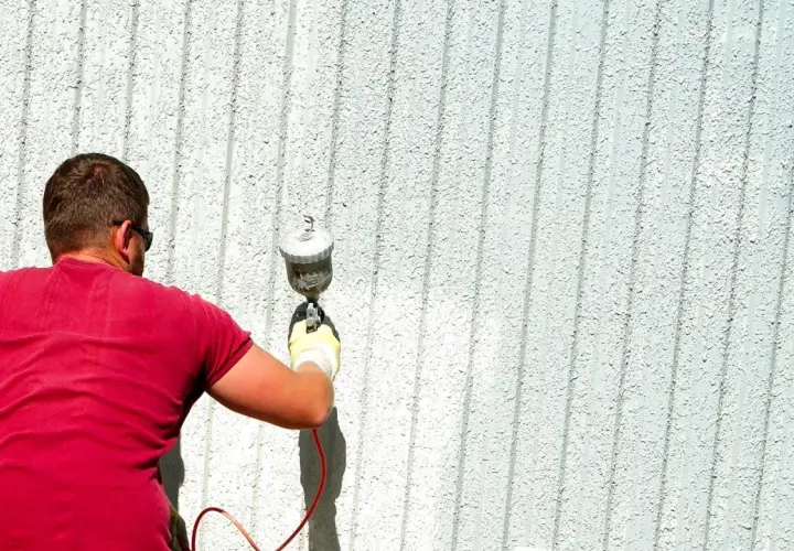 Uomo con maglietta rossa effettua verniciatura a spruzzo fai da te su una parete esterna bianca e rigata.