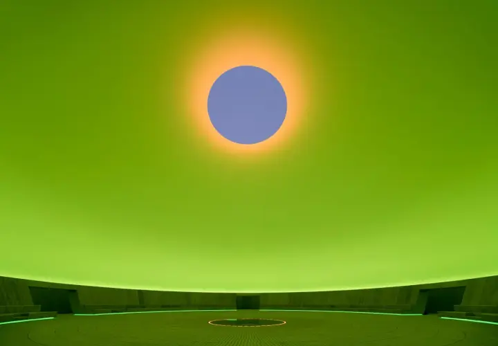 Interno di un'installazione di James Turrell: un cerchio blu su sfondo verde brillante, con un alone arancione. La sala circolare ha sedute e un cerchio luminoso al centro.