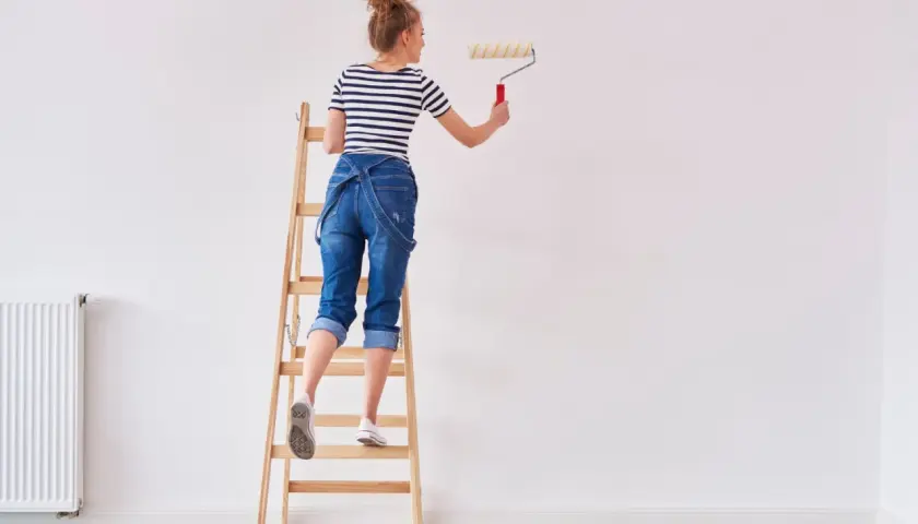 Donna su scala per pitturare casa fai da te, con rullo in mano.