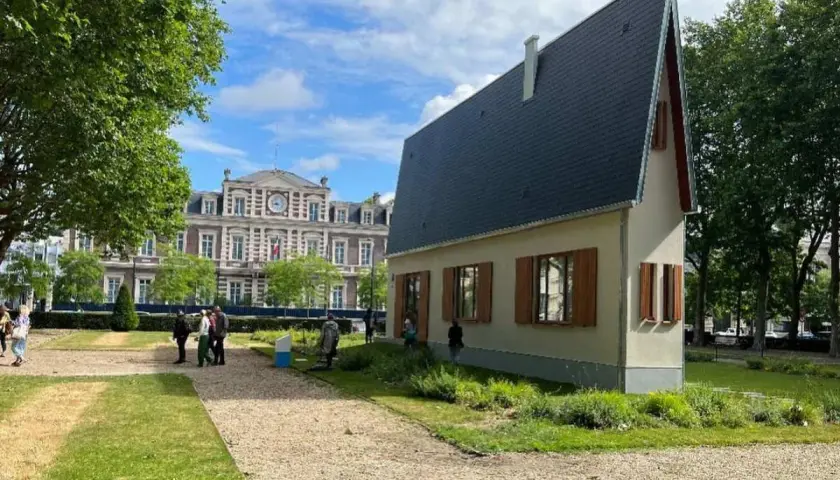 La casa più piccola del mondo, un'abitazione compatta con tetto spiovente, si erge in un parco con un edificio storico sullo sfondo.