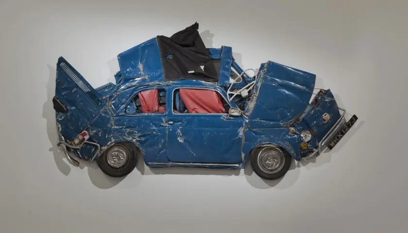 Opera di Ron Arad: una Fiat 500 blu, schiacciata e deformata, con interni rossi, appesa a un muro.
