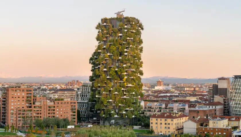 Il Bosco Verticale di Milano, un grattacielo ricoperto di alberi e piante, si staglia contro un cielo al tramonto, circondato da altri edifici della città.
