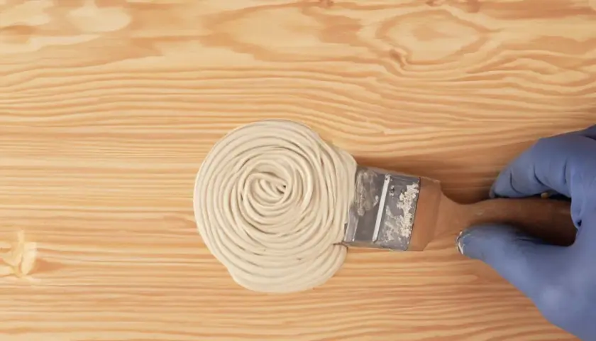 Mano con guanto blu applica stucco con pennello su legno. Guida pratica su come stuccare il legno per una finitura perfetta.