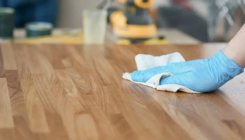 Mano con guanto blu che pulisce un tavolo di legno con un panno, mentre si valuta se sia meglio olio di lino crudo o cotto per la finitura.