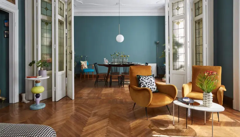 Soggiorno con poltrone ocra, tavolino moderno e un tavolo da pranzo in stile direttorio sullo sfondo. Porte a vetri decorati.