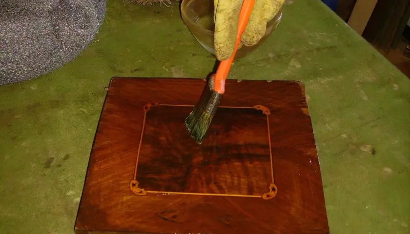 Guanto giallo applica liquido con pennello arancione su un mobile in legno scuro, per imparare come sverniciare un mobile.