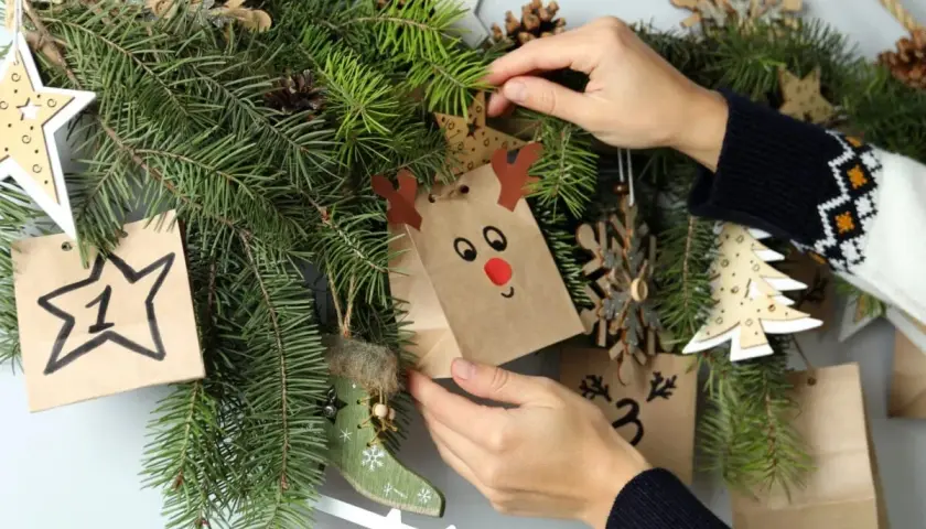 Mani che decorano un calendario dell'avvento con sacchetti natalizi fai da te, pigne e decorazioni in legno.
