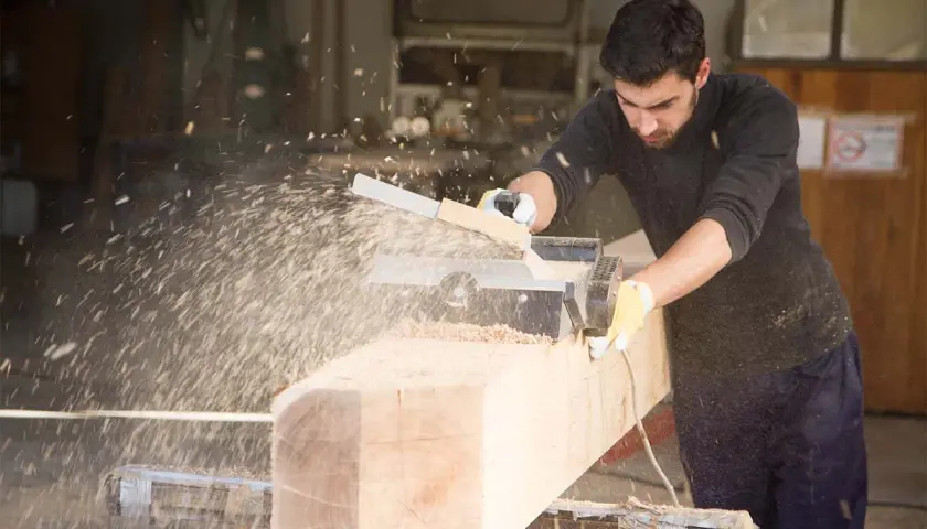 Un falegname usa una piallatrice per levigare un grosso tronco di legno, creando una nuvola di segatura. Il piallare significa rendere liscio e uniforme.