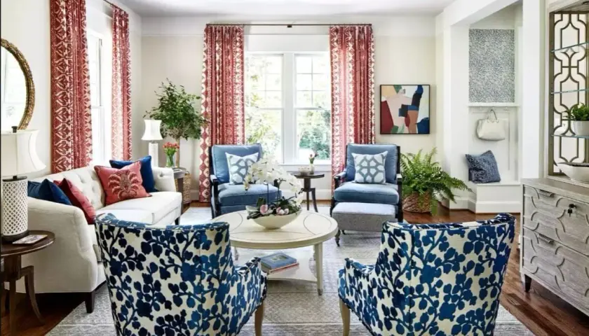Salotto in stile grandmillennial con divano bianco, poltrone blu e sedie con fantasia floreale blu e bianca. Tende rosse e specchio dorato.