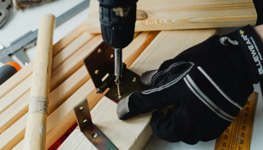 Mano guantata usa un avvitatore per unire due pezzi di legno con una staffa angolare e viti, mostrando come unire due pezzi di legno con viti.