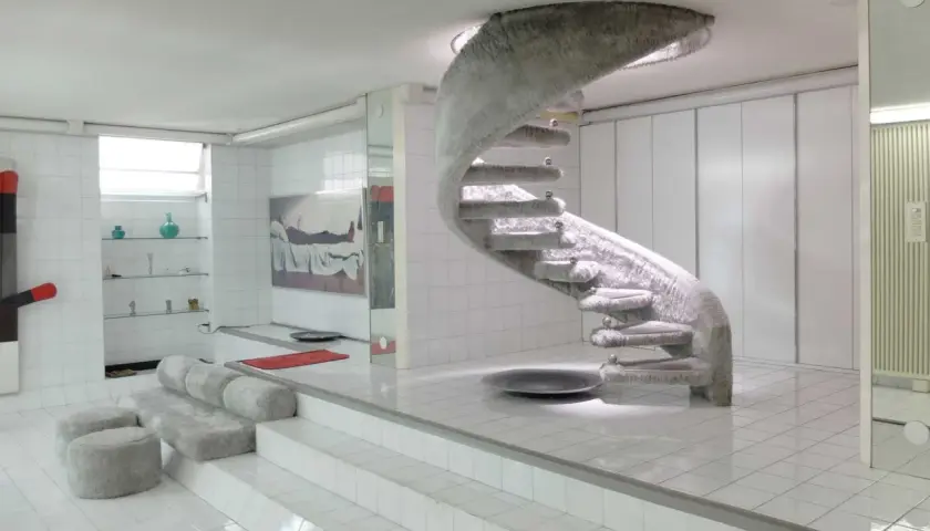 Interni futuristici con scala a chiocciola rivestita in pelliccia, un omaggio a Nanda Vigo. Divani e pouf morbidi completano l'ambiente.