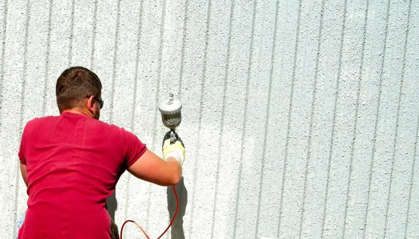 Uomo con maglietta rossa effettua verniciatura a spruzzo fai da te su una parete esterna bianca e rigata.