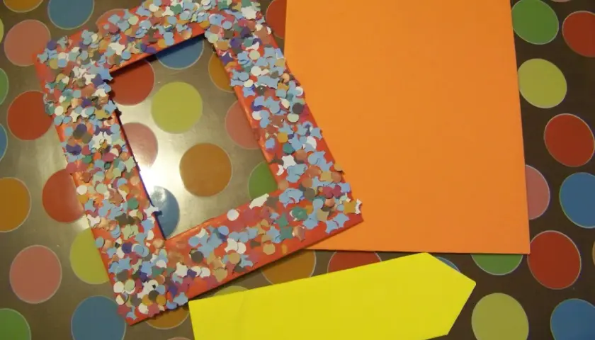 Cornice portafoto fai da te decorata con coriandoli colorati, accanto a fogli arancioni e gialli.