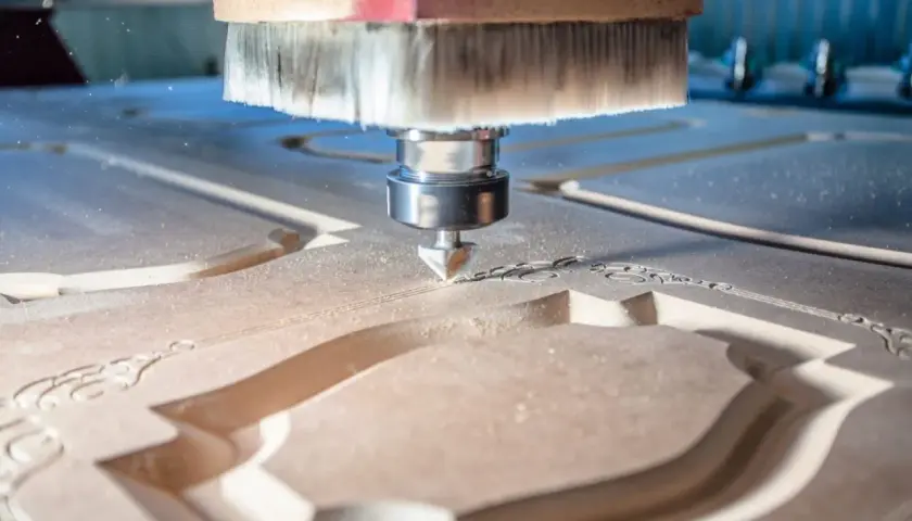 La fresa CNC sta lavorando il legno, creando un intricato motivo decorativo. La polvere di legno vola mentre la macchina esegue il suo lavoro di precisione.