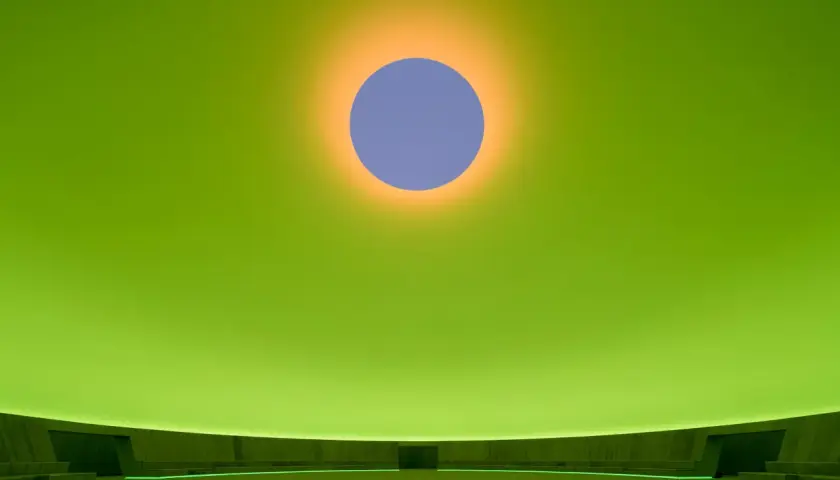 Interno di un'installazione di James Turrell: un cerchio blu su sfondo verde brillante, con un alone arancione. La sala circolare ha sedute e un cerchio luminoso al centro.