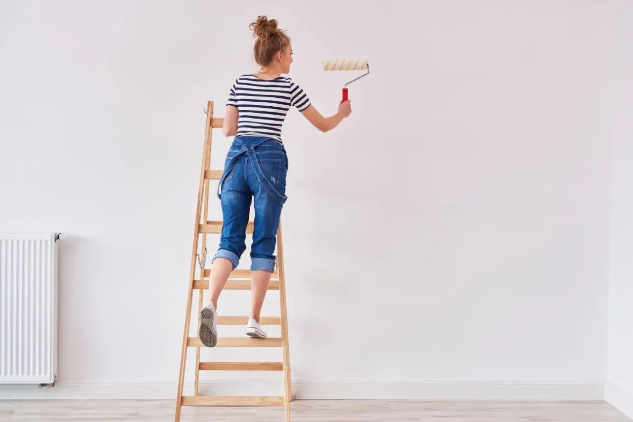 Donna su scala per pitturare casa fai da te, con rullo in mano.