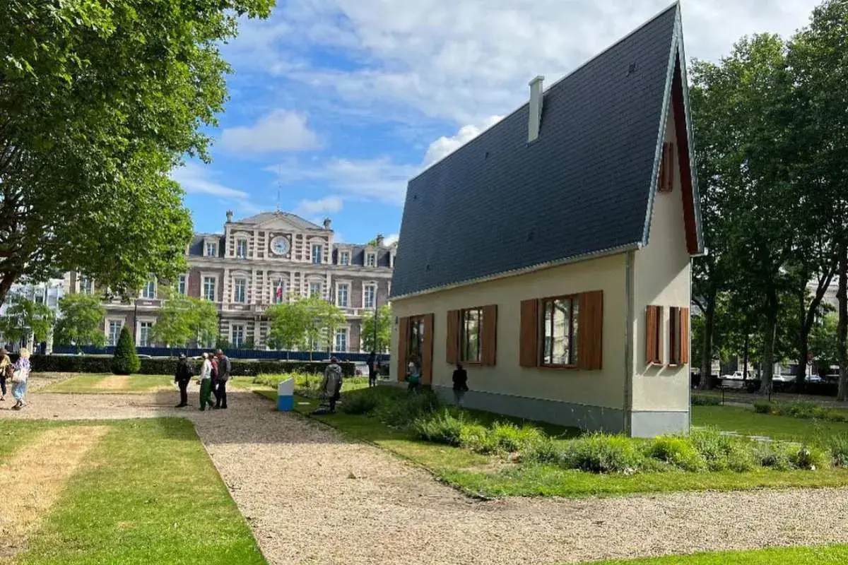 La casa più piccola del mondo, un'abitazione compatta con tetto spiovente, si erge in un parco con un edificio storico sullo sfondo.