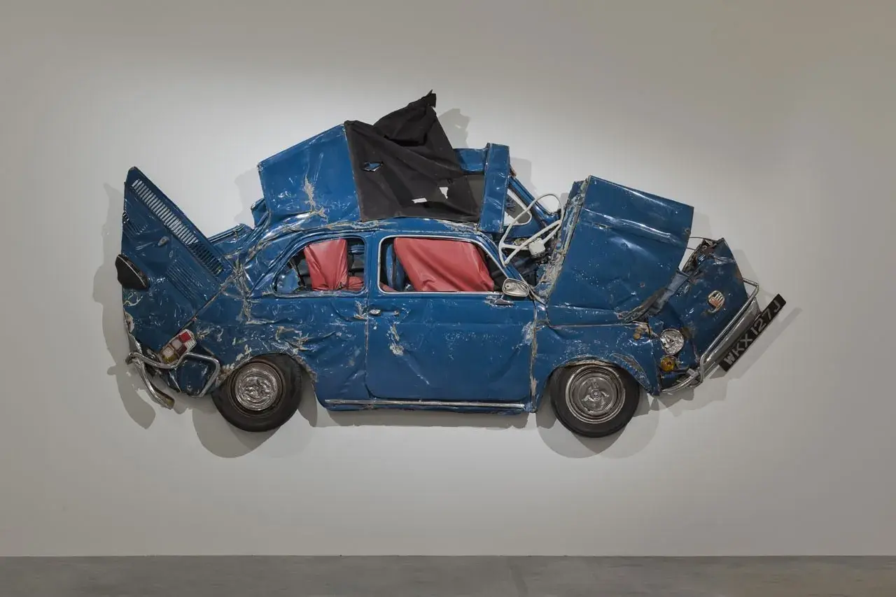 Opera di Ron Arad: una Fiat 500 blu, schiacciata e deformata, con interni rossi, appesa a un muro.
