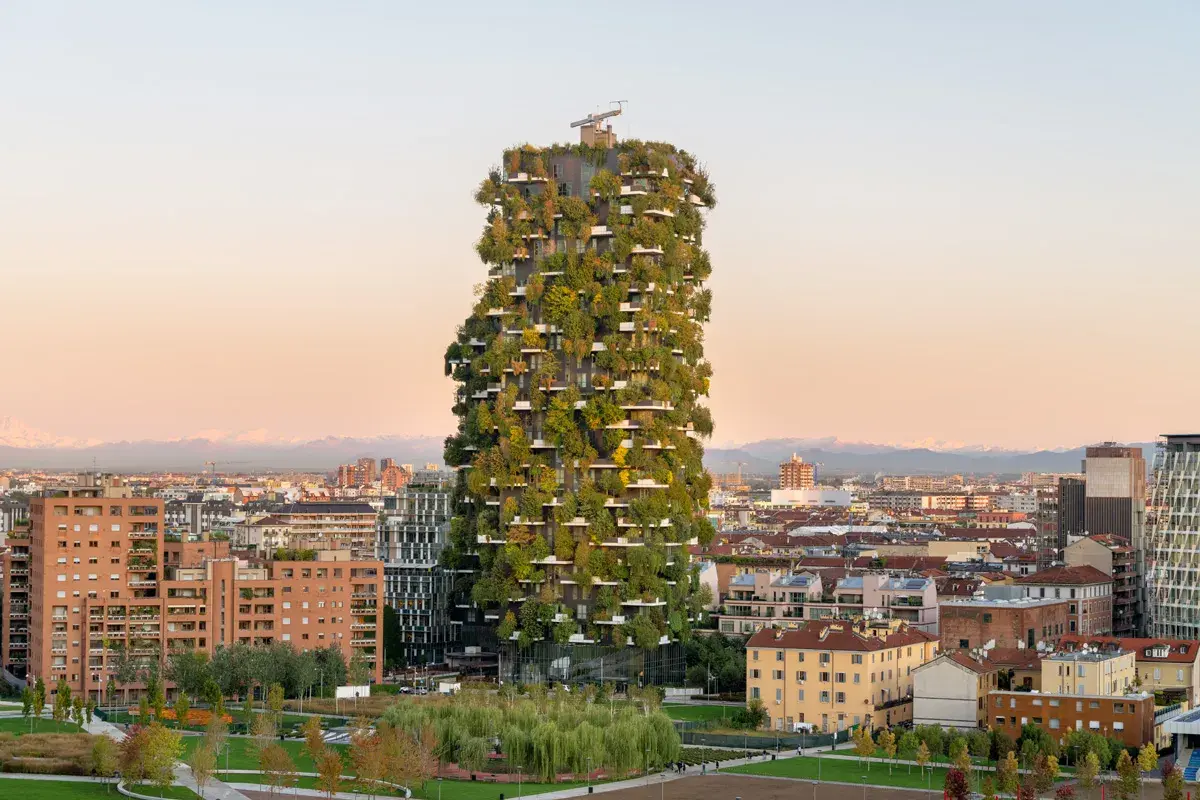Il Bosco Verticale di Milano, un grattacielo ricoperto di alberi e piante, si staglia contro un cielo al tramonto, circondato da altri edifici della città.